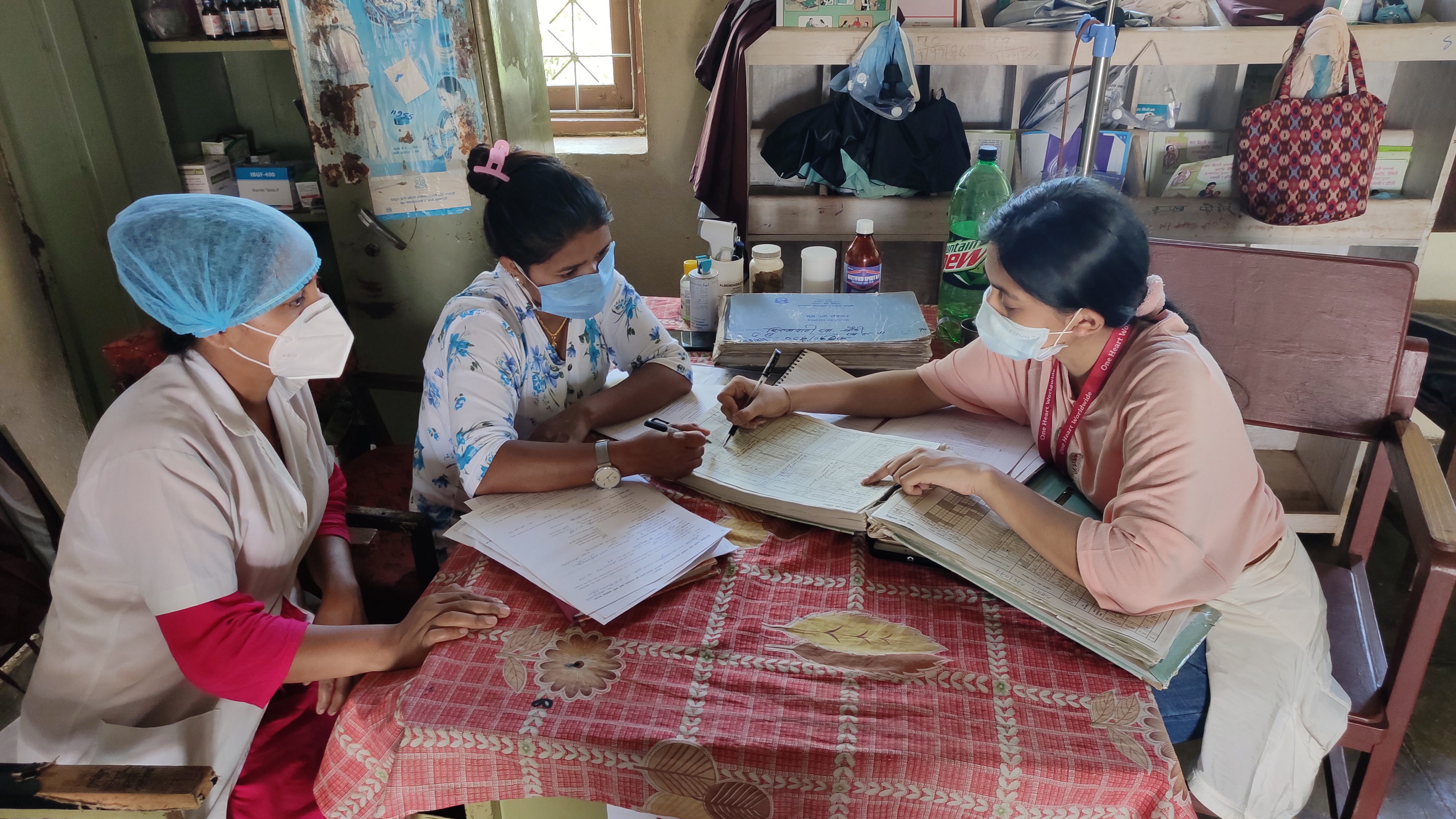 Credit: One Heart Worldwide. Description: Photo of three women sitting at a table for nursing staff training. One women is pointing to a book and the other two are looking at what she's pointing out.