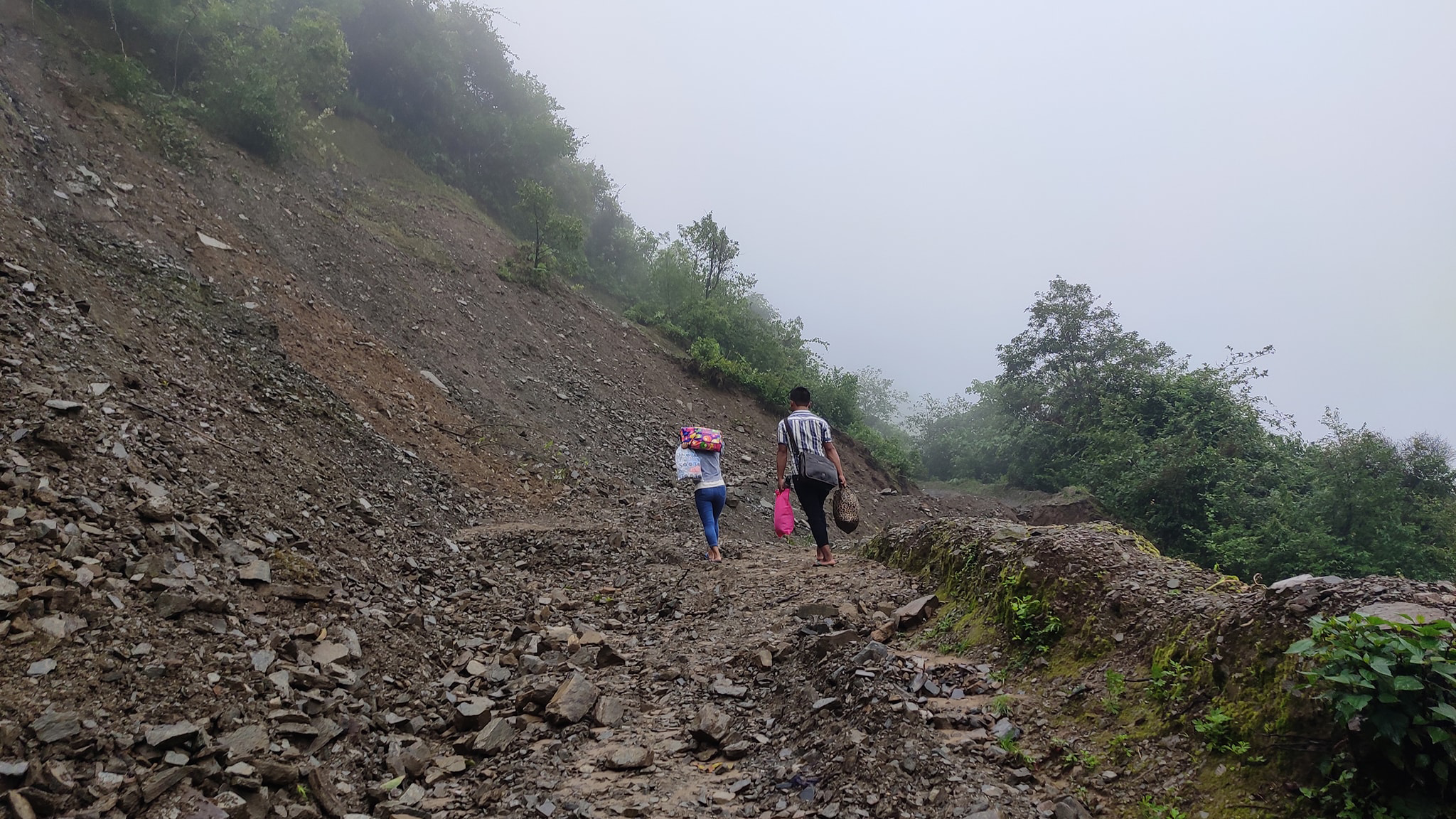 Credit: One Heart Worldwide. Description: Photo shows two people walking barefoot on rugged terrain. Between the two of them, they are carrying five bags.