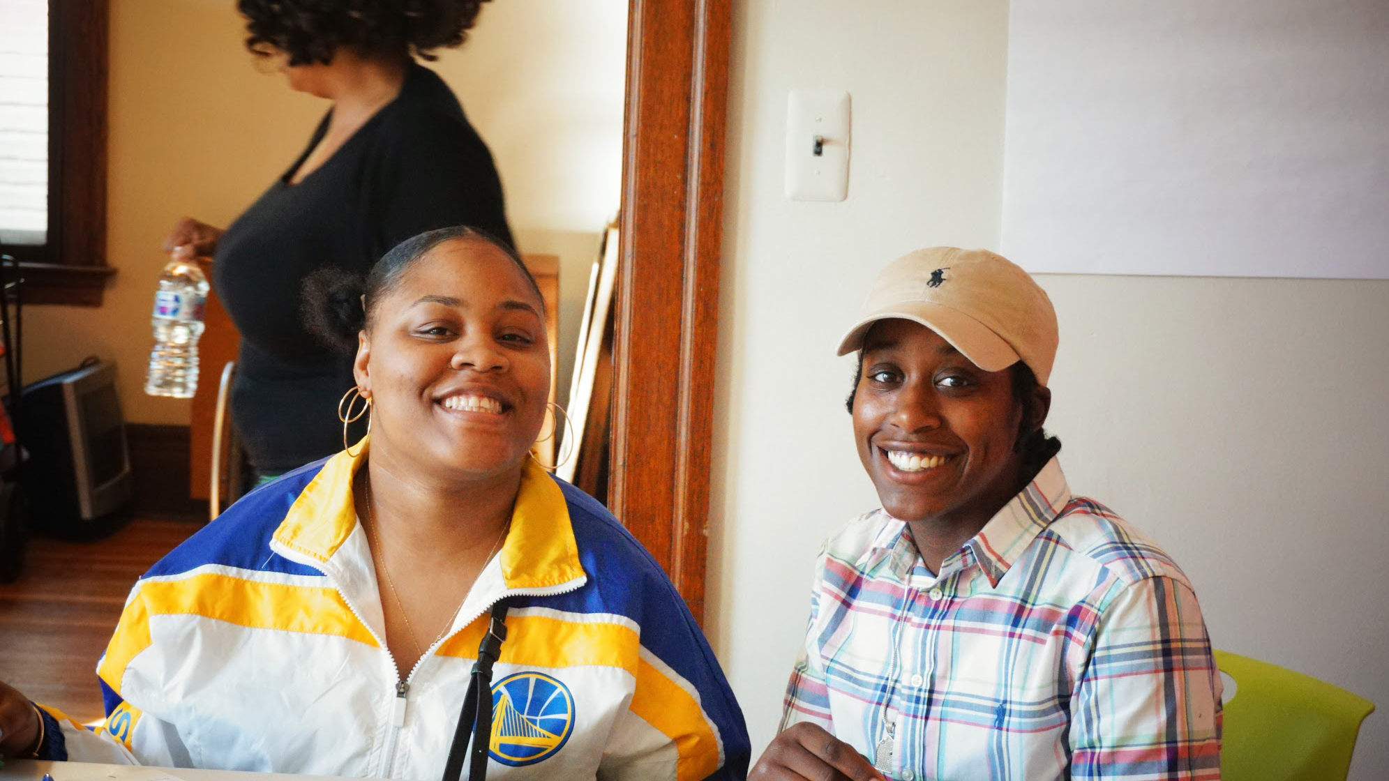 Photo shows two people sitting at a table. The one on the left is wearing a yellow, white and blue windbreaker. The one on the right is wearing a plaid shirt and a tan hat. Both are smiling.